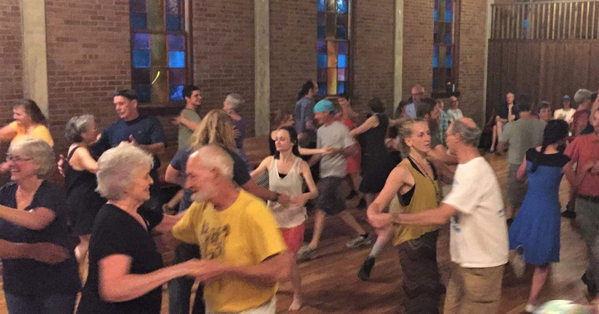 A crowded hall of dancers of all ages swinging partners on a wooden floor